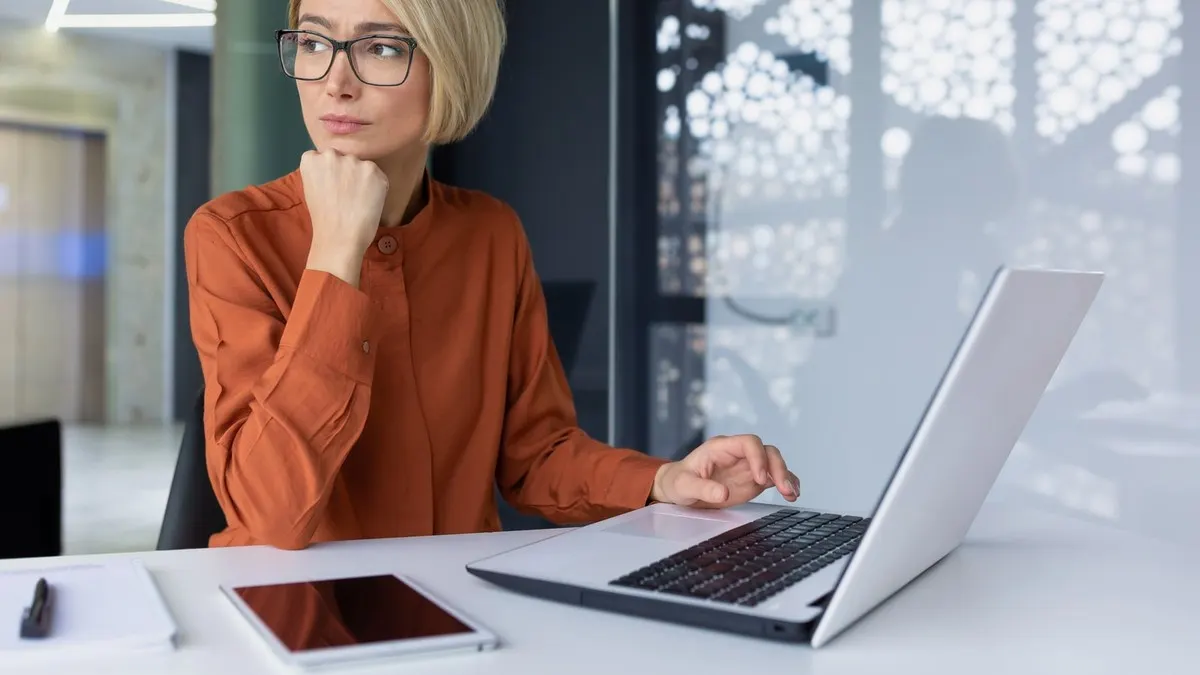 A woman in glasses and an orange blouse rests her chin on her fist and looks away from her laptop, weighing a decision.