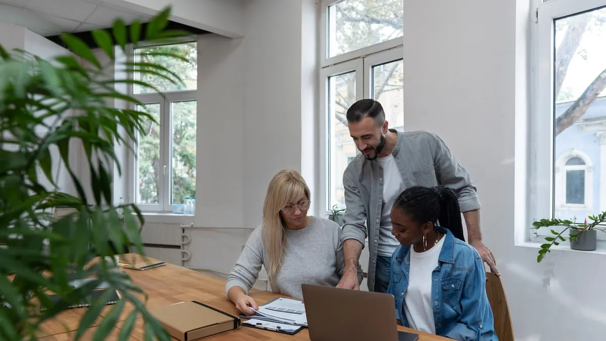 Three colleagues review papers and a laptop together at a wooden table in a bright office with plants