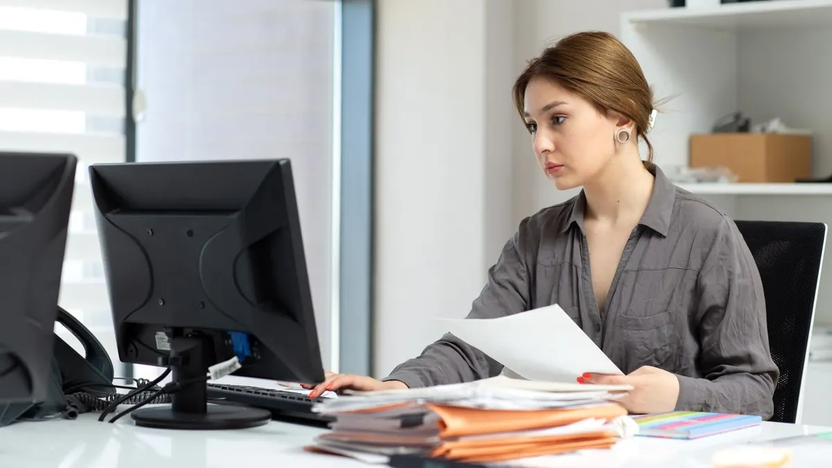 A woman reading a printed document beside a stack of files at her desktop computer in a bright white office