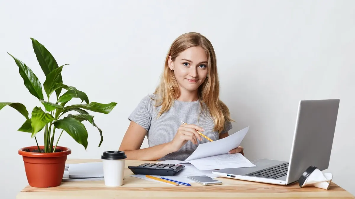 A woman at a white desk working through papers with a calculator and an open laptop
