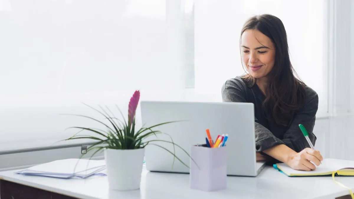 A smiling woman writes in a notebook beside her laptop at a white desk by a bright window