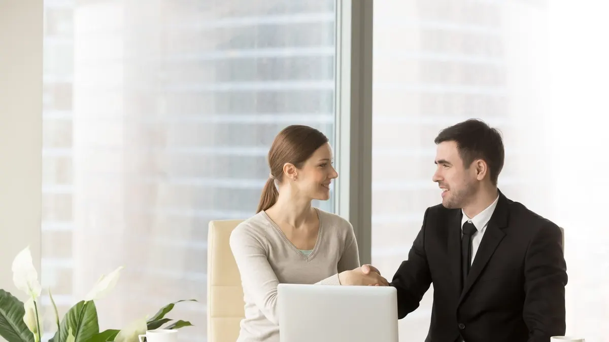 A woman and a man in a suit shake hands across a desk with a laptop by a tall office window