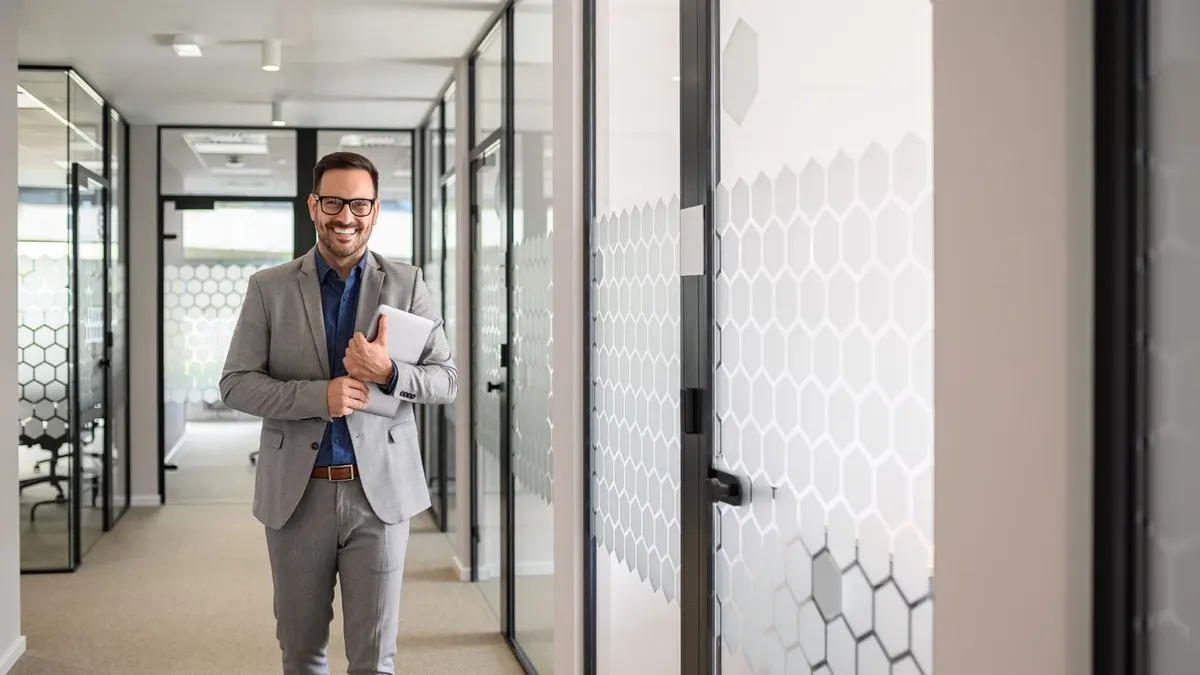 A smiling professional in glasses walking down a bright glass-walled office corridor with a laptop under his arm