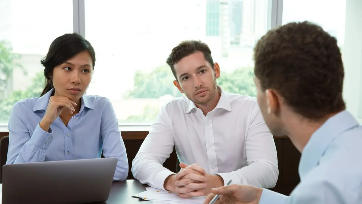 Two interviewers at a table listen closely to a candidate seated across from them by a bright window