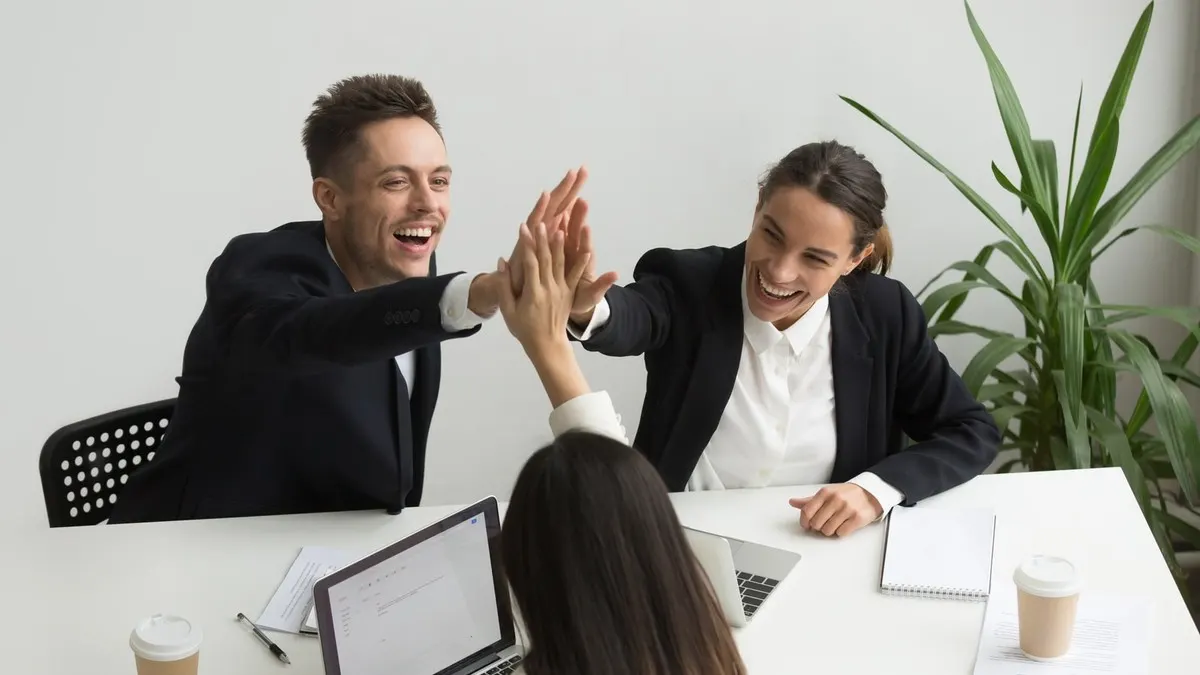Three laughing colleagues share a high five across a white meeting table with laptops and coffee