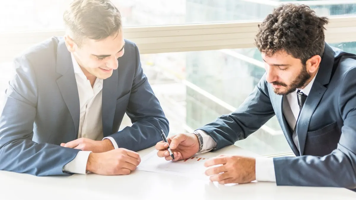 Two businessmen in suits at a white desk by a window, one of them signing a document