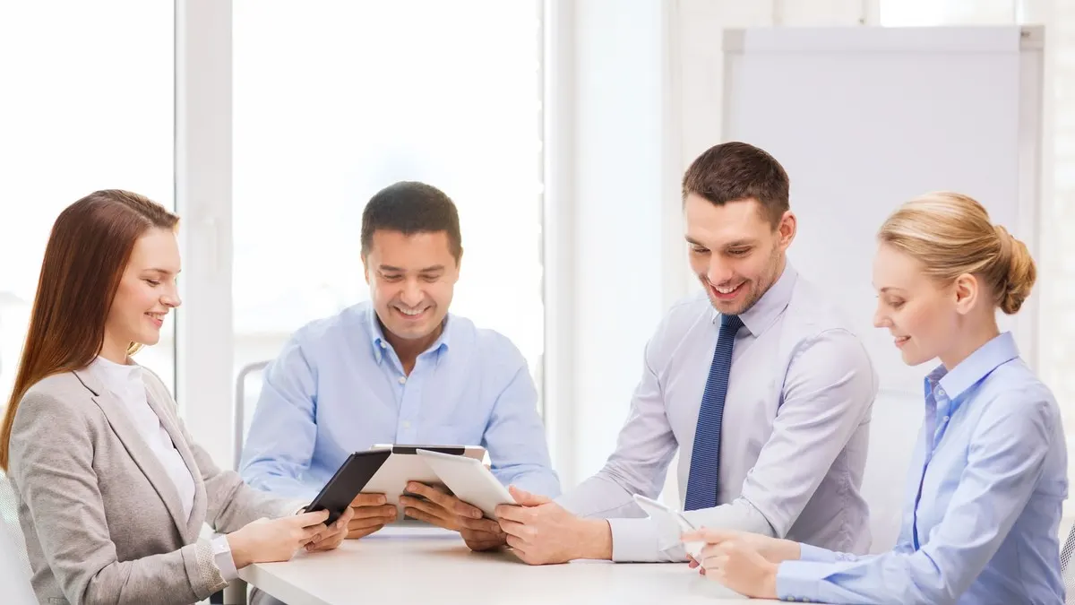 Four smiling colleagues gather around tablets at a table in a bright white office to work through a plan together.
