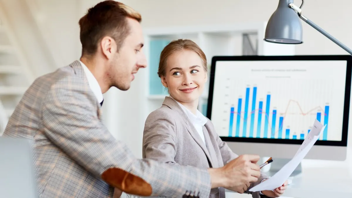 Two colleagues discuss a printed report in front of a monitor showing a bar chart