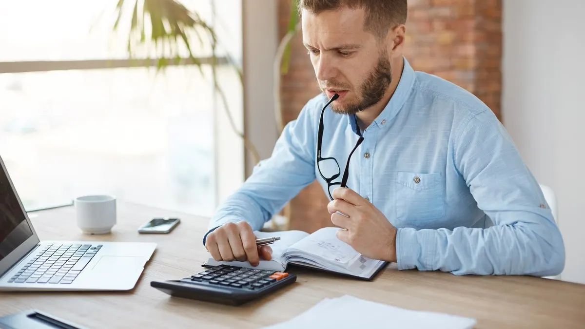 A man in a blue shirt working with a calculator and a notebook at a sunlit office desk