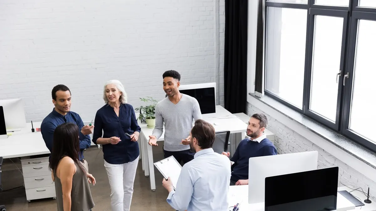 A senior woman leads an informal standing discussion with her team in a bright loft office with tall windows.