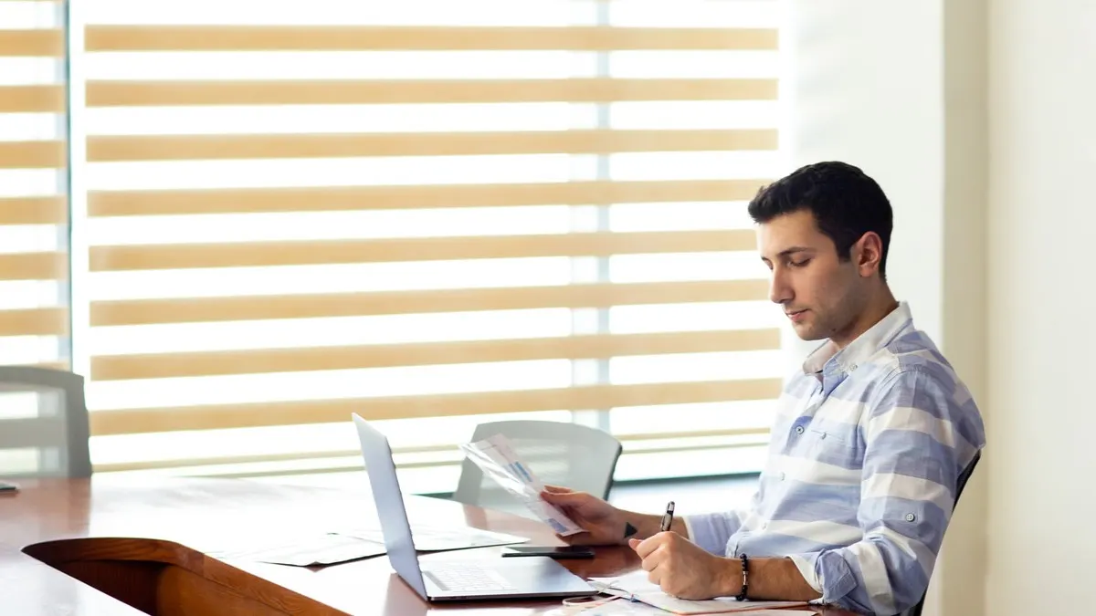 A man in a striped shirt reviewing papers beside his laptop at a long table in a sunlit room