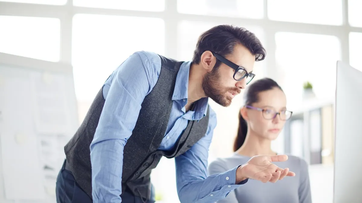 A man in glasses leans over to help a colleague with her work at a computer in a bright office