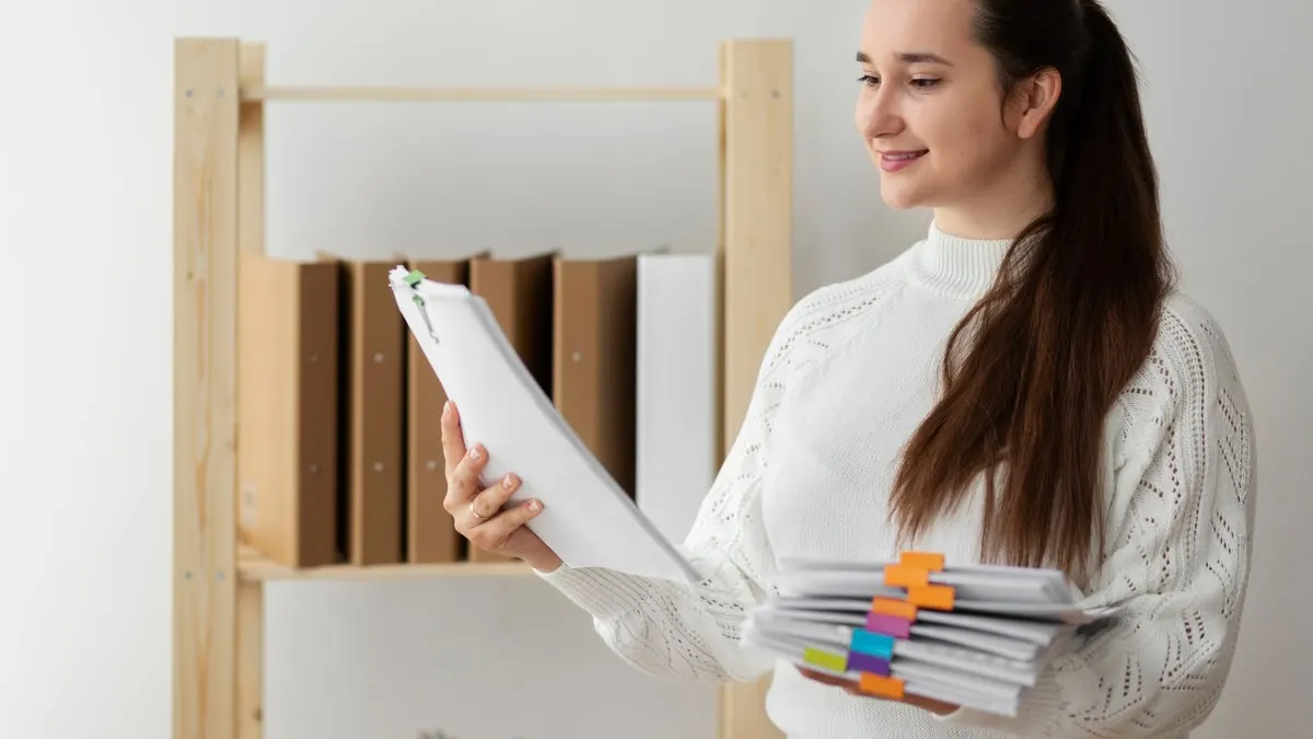 A young woman holding a stack of tabbed documents beside a shelf of binders in a bright office