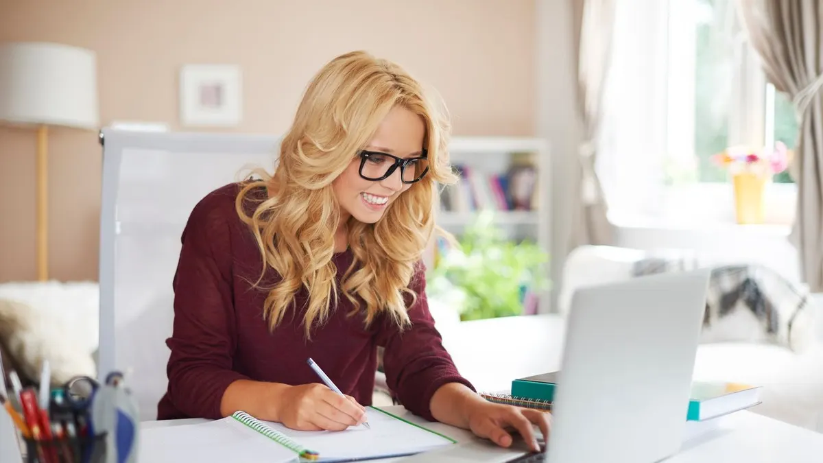 A smiling woman in glasses writes notes while working on a laptop at a desk in a bright room