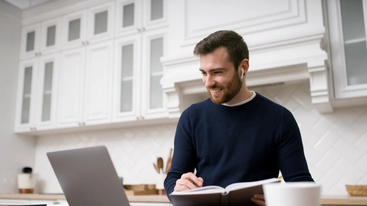 A smiling man studying from a book beside his laptop in a bright white kitchen