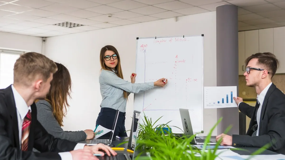 A woman draws on a flipchart while her colleagues around the table follow along in a bright meeting room.