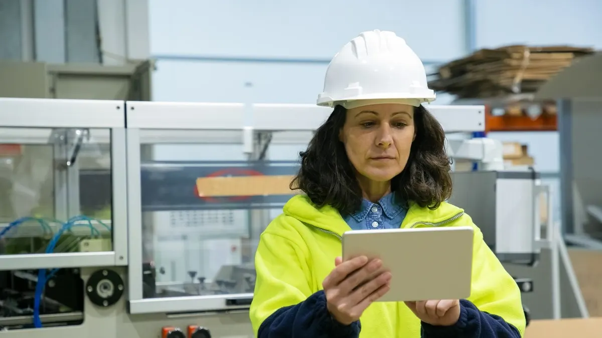 A woman in a white hard hat and safety jacket checks a tablet beside machinery in a factory