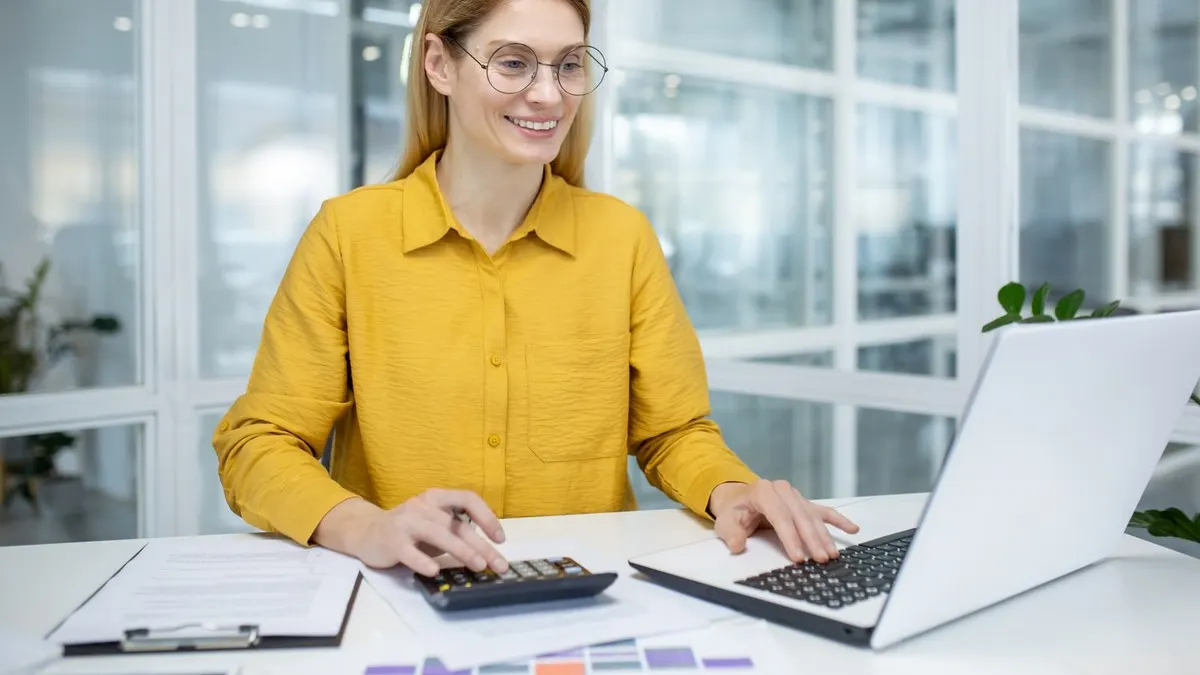 A smiling woman in a yellow shirt works out figures on a calculator beside her laptop in a bright office.
