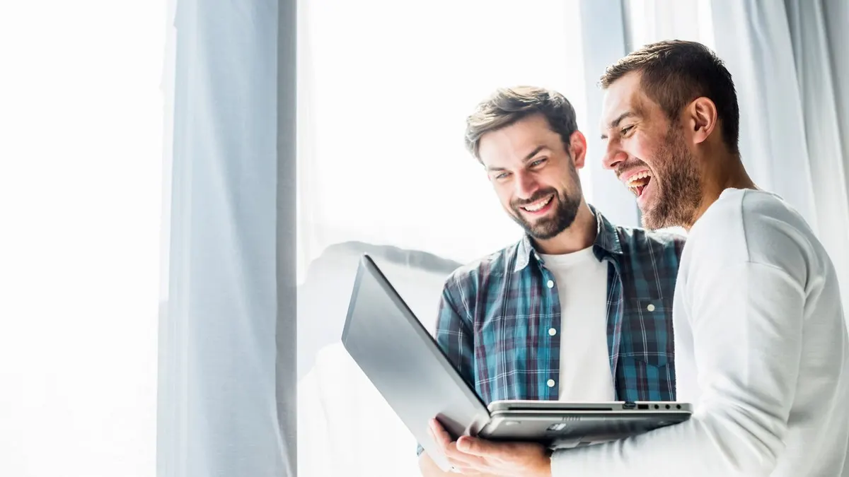 Two smiling men look at an open laptop together by a bright window