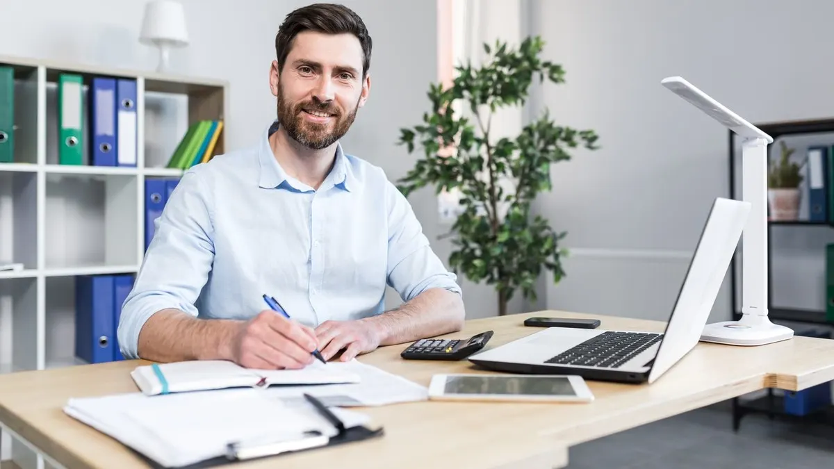 A smiling professional sits at his desk with a laptop and notes in a bright, modern office.