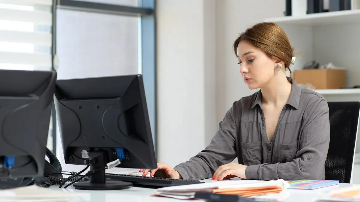 A woman in a grey shirt types at a desktop computer beside stacks of papers in a bright office