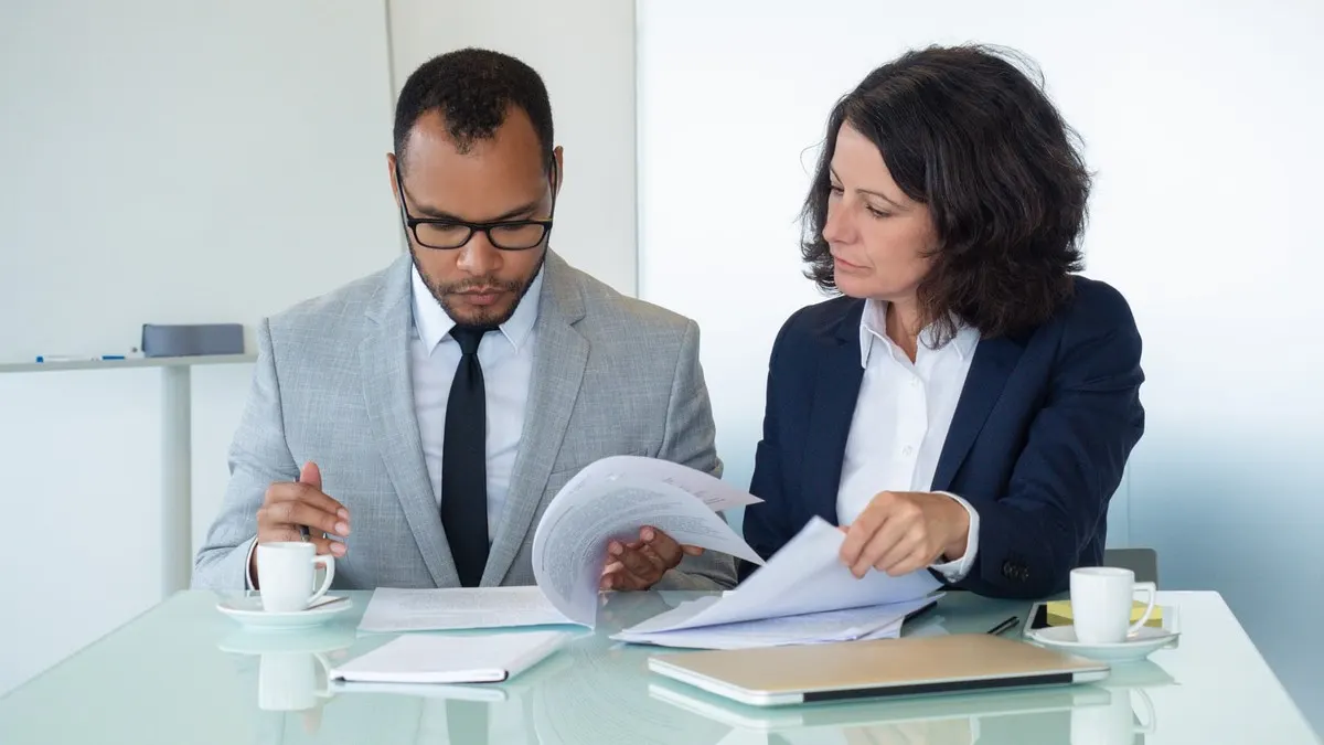 Two colleagues in suits review printed documents together at a glass table in a bright office