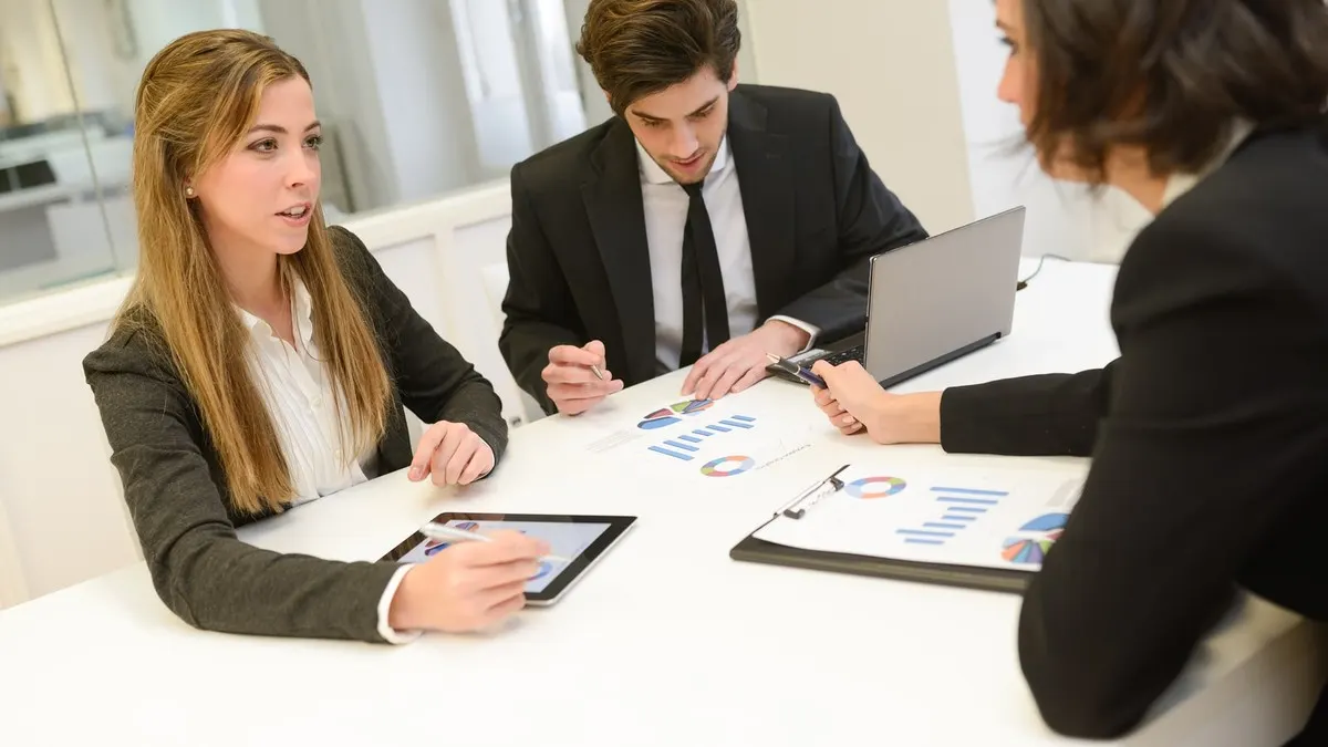 A business analyst with a tablet sharing her view with colleagues around a white table covered in printed charts