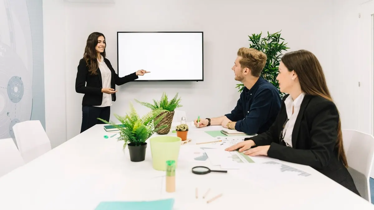 A businesswoman presenting at a wall screen to two business partners seated at a white table in a bright meeting room