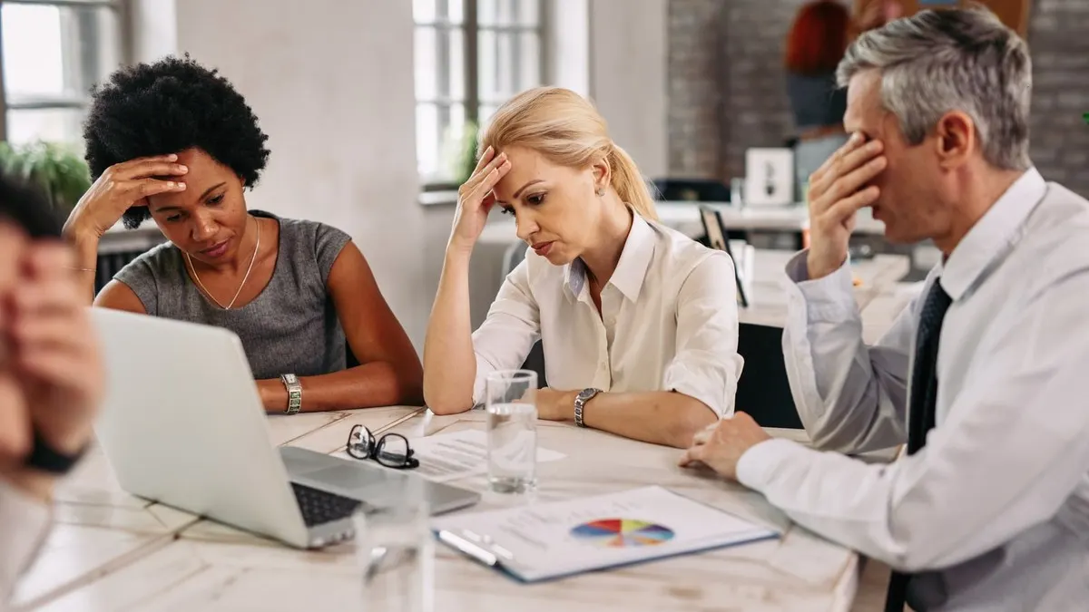 Three worried colleagues hold their heads while looking at a laptop and a report at a meeting table