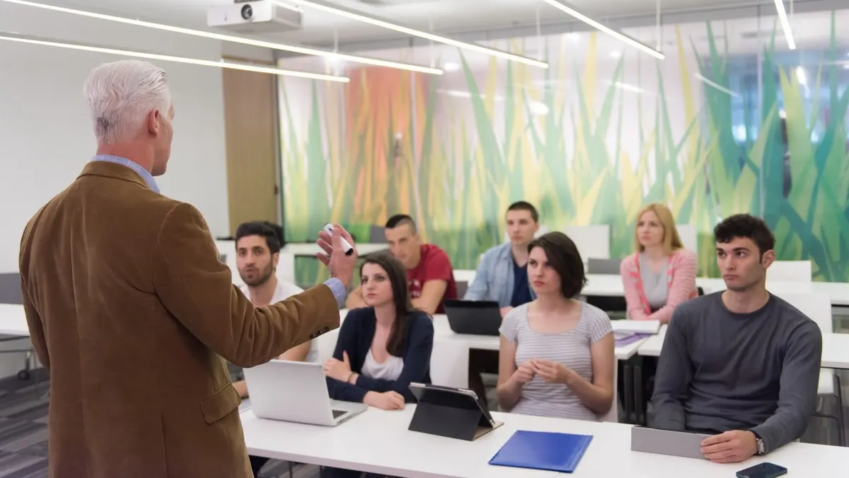 A grey-haired professor lecturing a class of adult students seated at laptops in a bright modern classroom