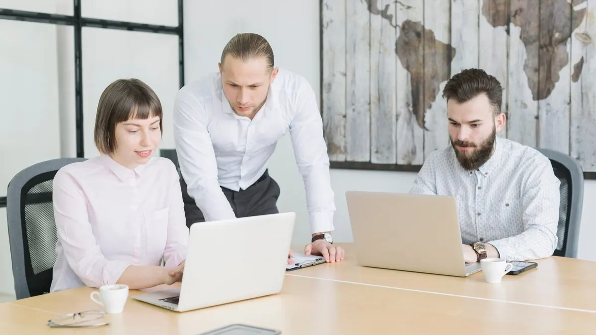 Three colleagues work on laptops at a shared table while one leans in to look at a screen