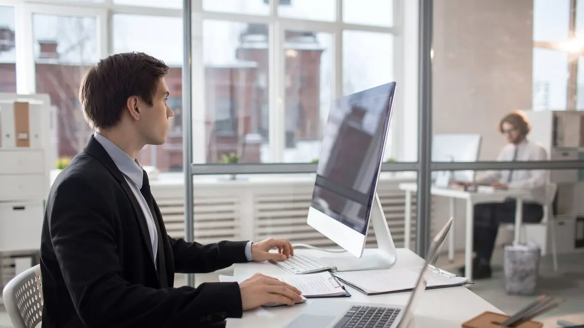 A man in a dark jacket working at a desktop computer in a bright glass-walled office