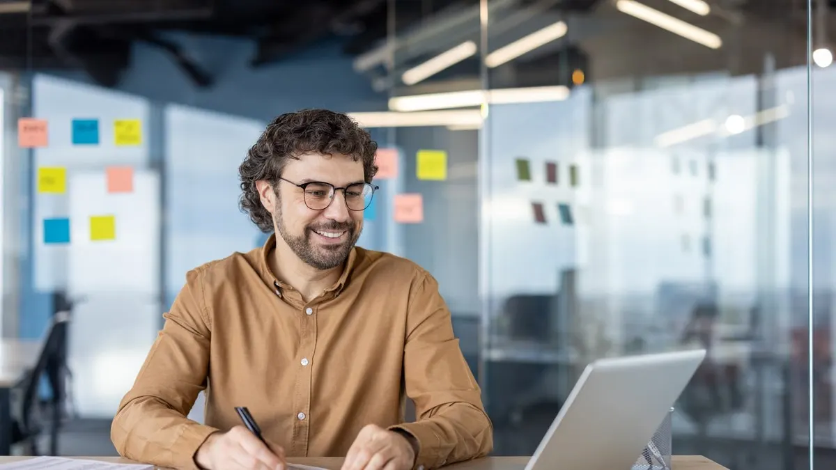 A smiling man in glasses takes notes beside his laptop in a glass-walled office with sticky notes on the wall.