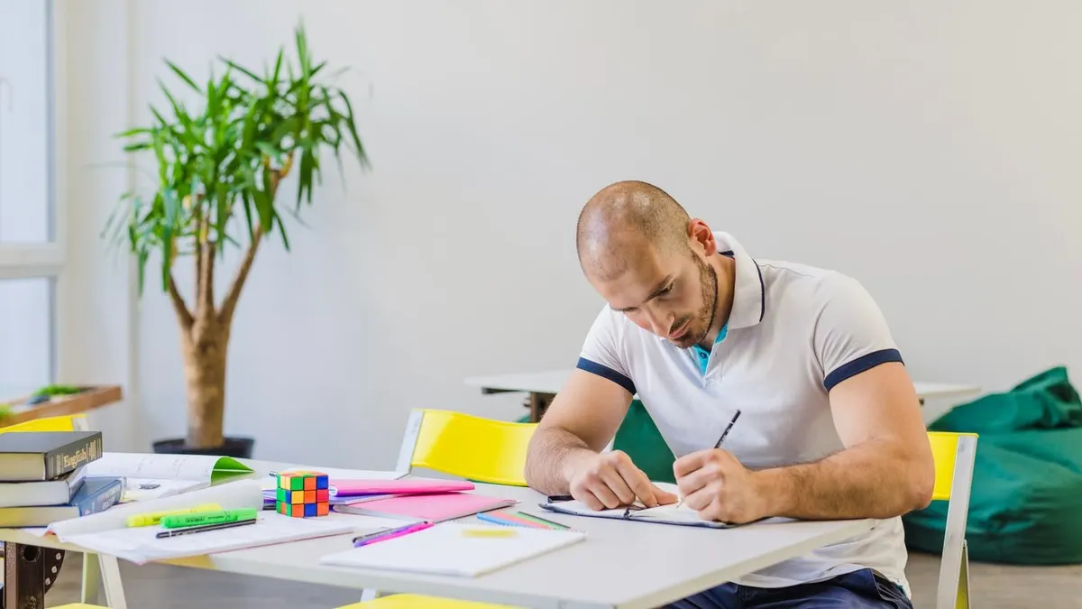 A man writes in a notebook at a white table spread with study books, highlighters and a puzzle cube.