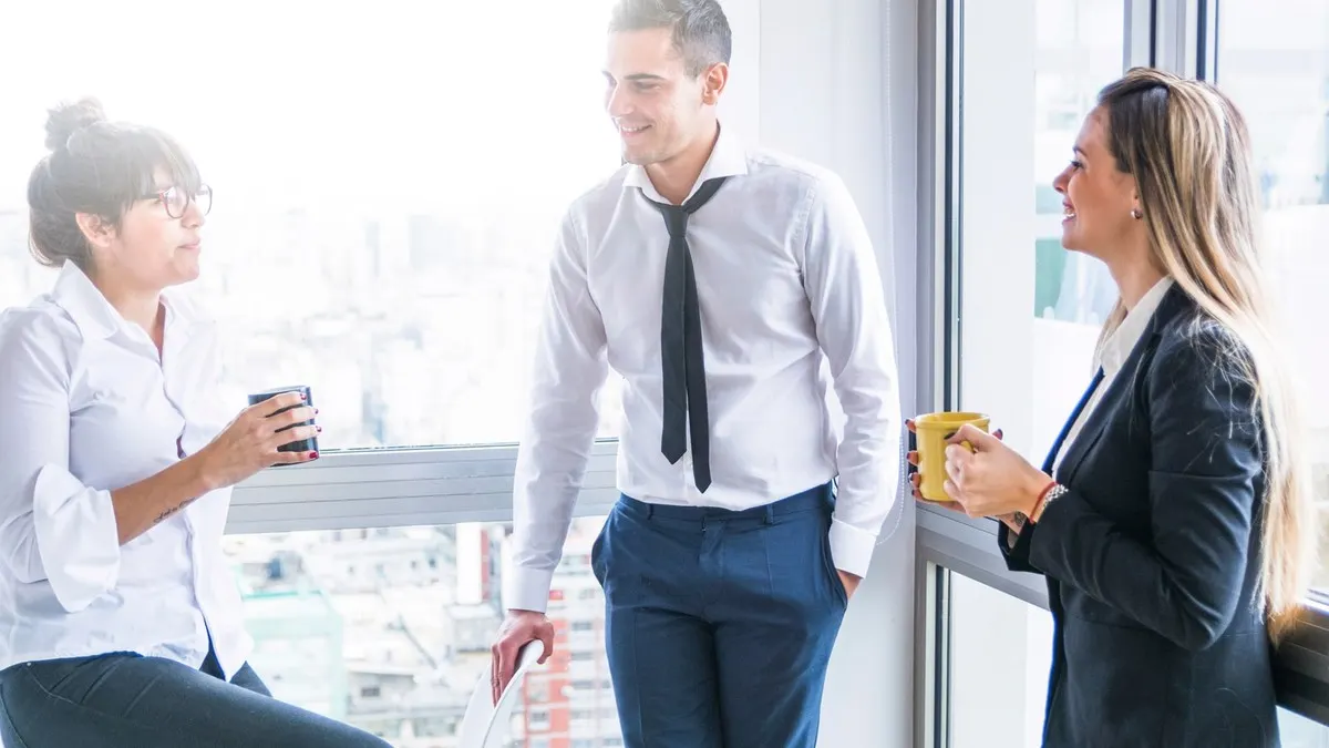 Three colleagues holding coffee cups and talking together beside a bright office window
