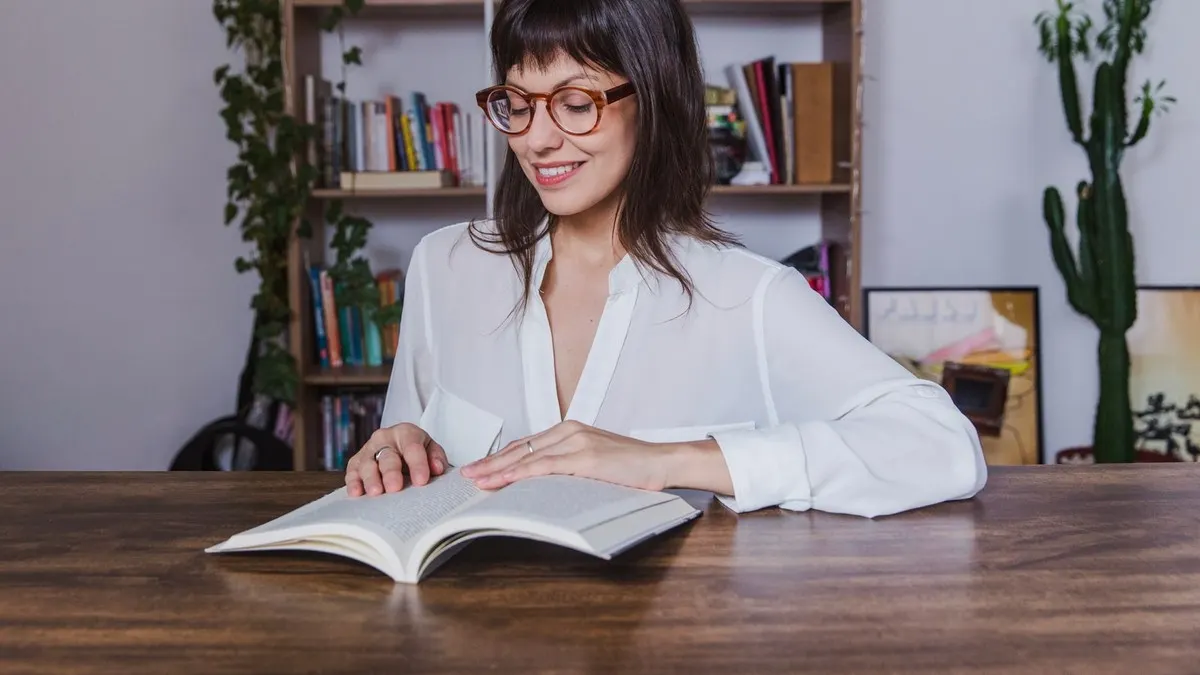 A smiling woman in glasses reads an open book at a wooden table in front of a bookshelf