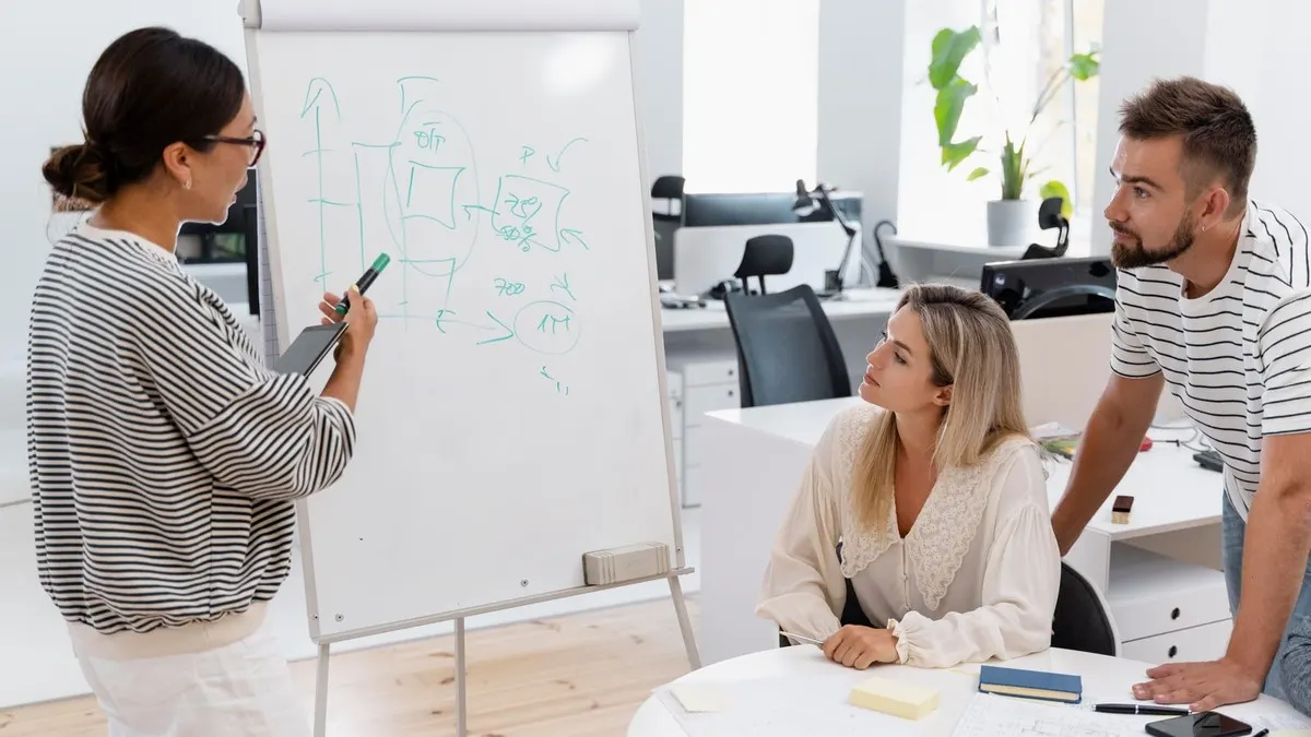 A woman sketching a diagram on a flip chart while two colleagues at a table weigh up the options in a bright office
