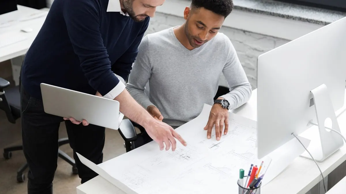 Two colleagues reviewing a printed plan together at a white desk beside a computer