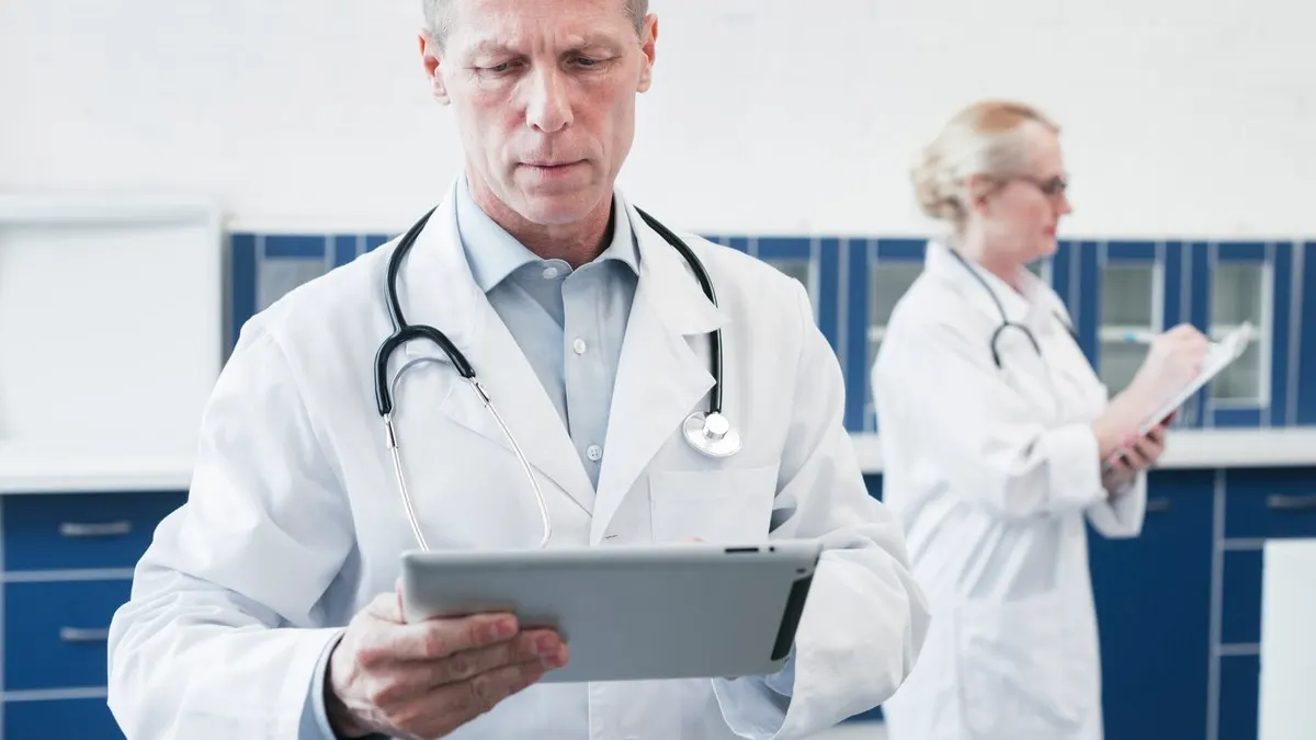 A doctor in a white coat reads a tablet while a colleague takes notes on a clipboard behind him