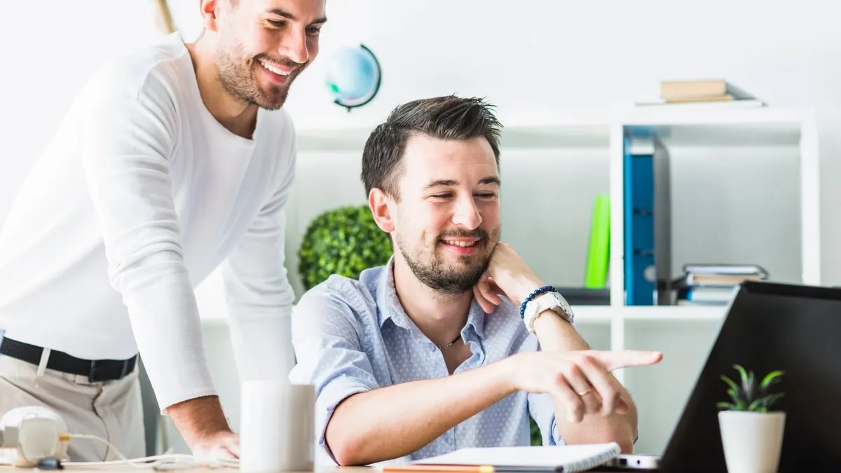 Two smiling colleagues looking at a screen together, one standing and one seated pointing at the work, in a bright white office