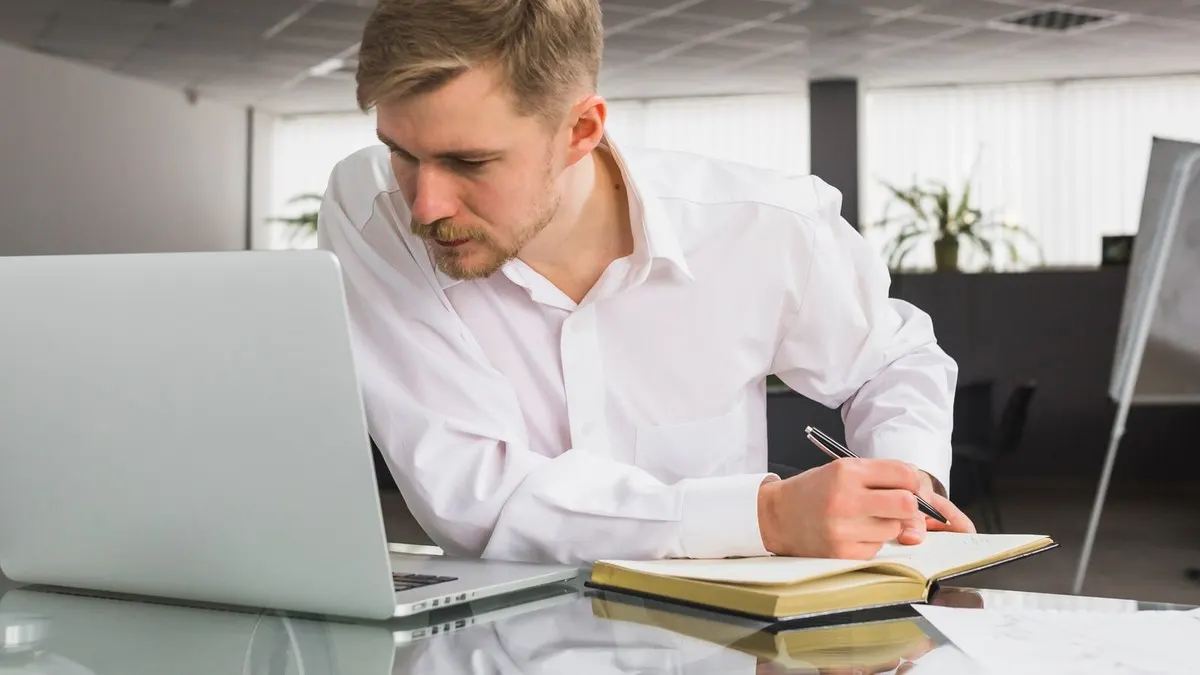A man in a white shirt takes notes in a notebook while reading from a laptop at an office desk