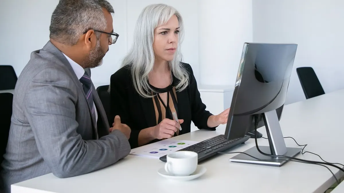 A man and a woman reviewing work on a computer monitor at a white desk in a bright office
