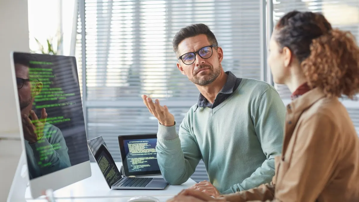 Two software developers discuss code shown on their computer screens at a desk in a bright office.