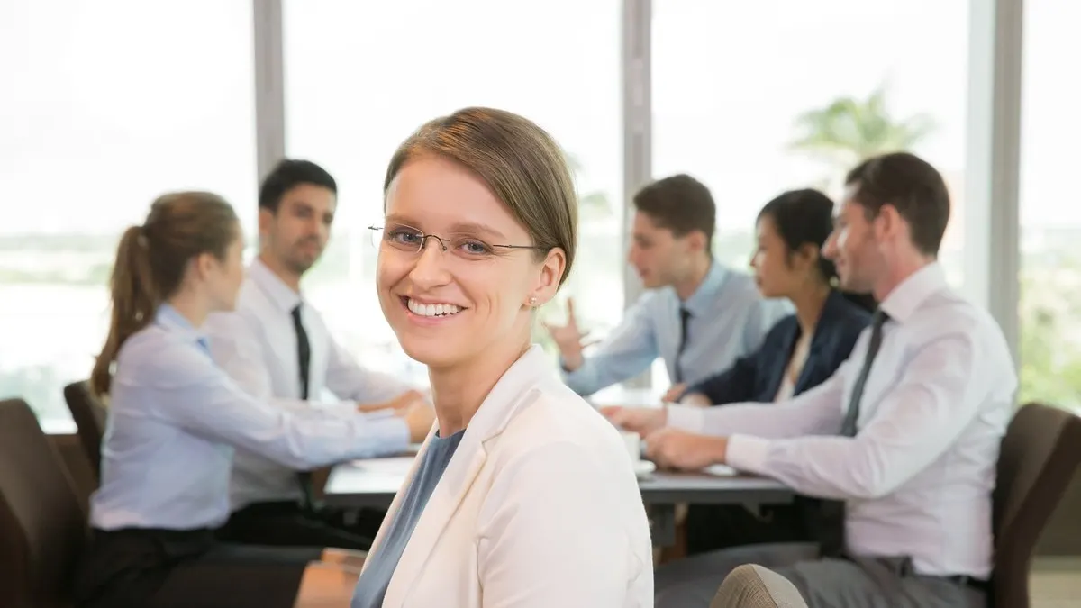 A smiling businesswoman in glasses in the foreground while her team meets around a table in a bright office