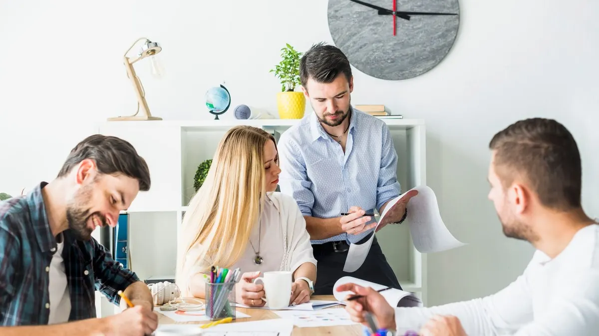 A busy team working together at a shared desk under a large wall clock in a bright white office