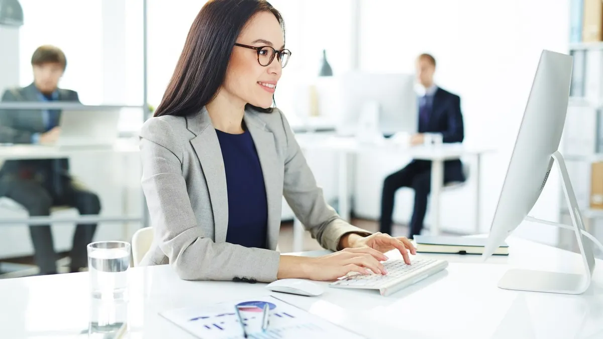 A smiling woman in glasses types at a desktop computer with a printed chart beside her in a bright office