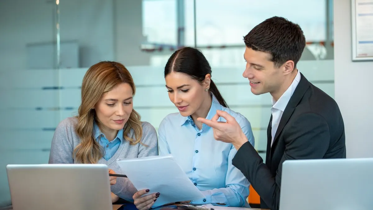 Three colleagues go through a printed document together between their laptops in a bright office.