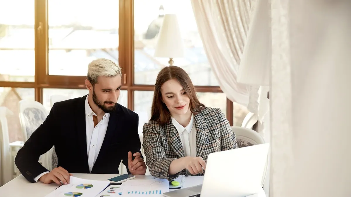 A man and a woman review printed charts and a laptop together at a table in a bright room with tall windows.
