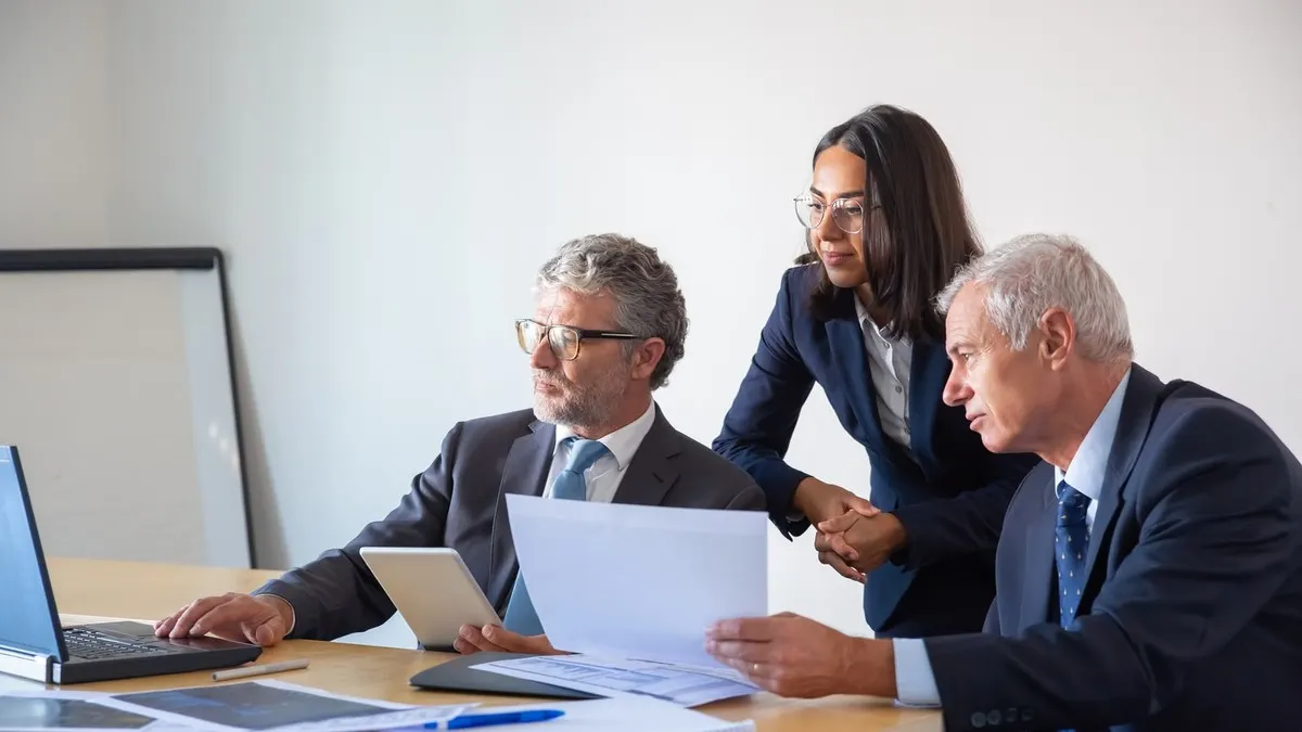 Two senior executives and a younger colleague in suits review papers and a laptop together at a meeting table.