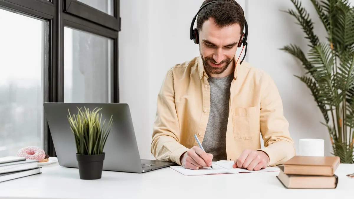 A smiling man wearing a headset takes notes beside his laptop at a white desk by a window